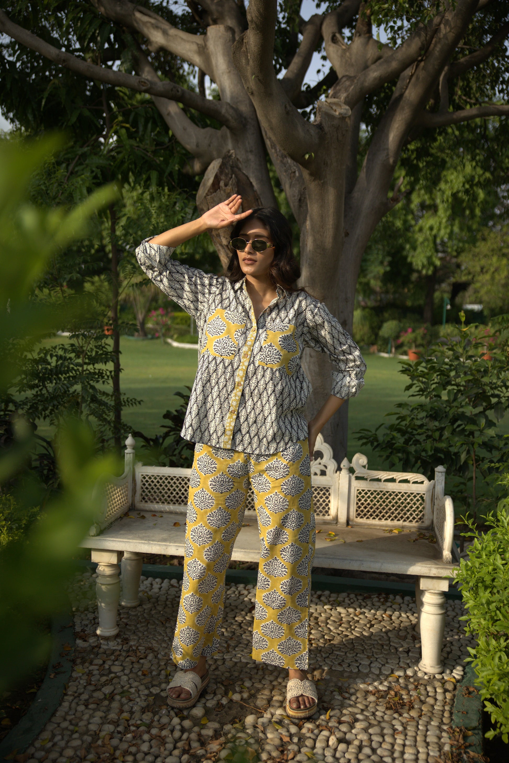Person in a patterned outfit standing in a garden with trees and a stone wall.